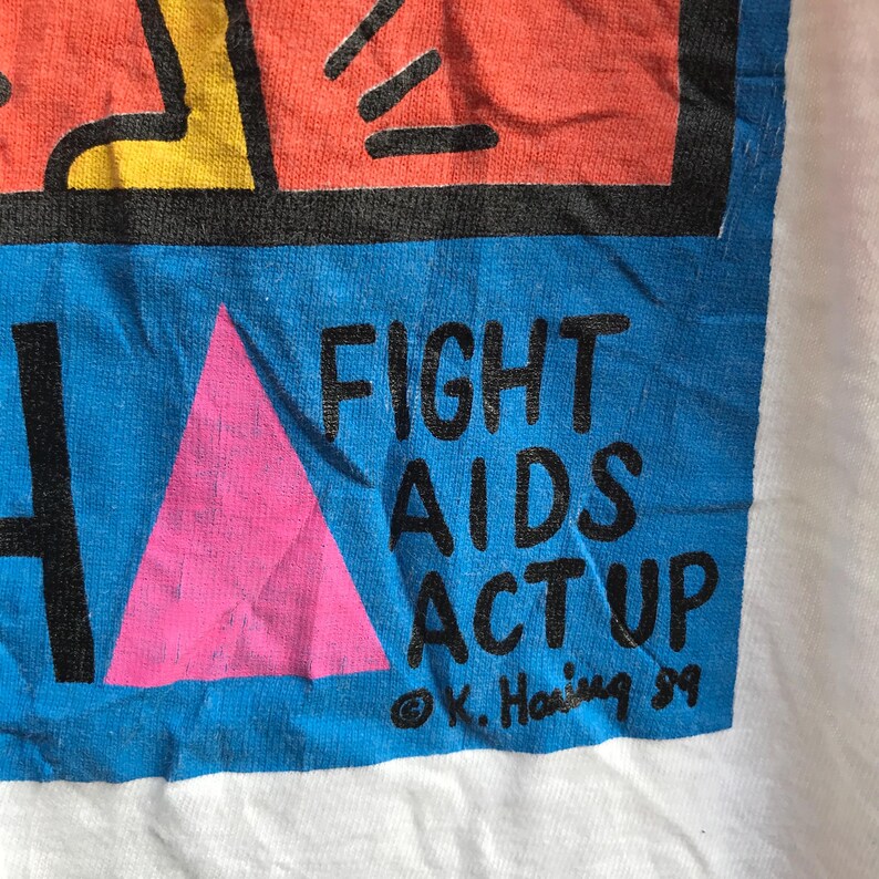 Pu&ograve; includere: Una maglietta bianca con una grafica blu e rosa. La grafica presenta un triangolo rosa, una figura nera e il testo "FIGHT AIDS ACT UP" con una firma che recita "&copy; K. Haring 89".