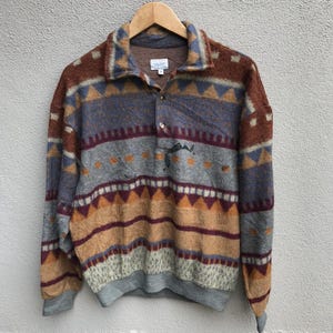 Op de afbeelding: Een vintage pullover met kraag en knoopsluiting. De trui heeft een geometrisch patroon in bruin, blauw, grijs en oranje tinten. De trui met lange mouwen heeft een relaxte pasvorm en geribde zoom en manchetten.