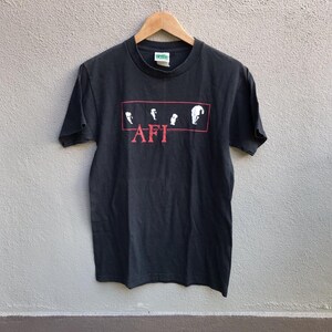 May include: Black t-shirt with a red rectangular graphic featuring four white silhouettes and the red text "AFI". The shirt is hanging on a wooden hanger against a neutral background.