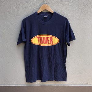 Peut inclure: T-shirt bleu marine avec un motif ovale jaune et rouge portant l'inscription "TOWER". Le t-shirt est suspendu à un cintre en bois. Le tissu semble doux.