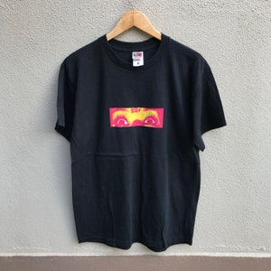 以下が含まれることがあります： 赤い瞳と黄色い瞼の目のグラフィックがプリントされた黒いTシャツ