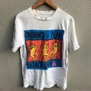 Pu&ograve; includere: Maglietta bianca con una stampa grafica di tre figure con le mani sulle orecchie. La grafica dice "Ignorance = Fear" e "Silence = Death". La grafica include anche il testo "Fight AIDS Act Up".