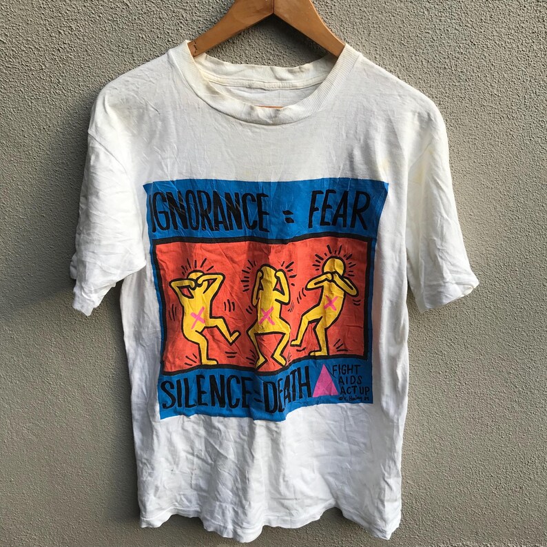 Pu&ograve; includere: Maglietta bianca con una stampa grafica di tre figure con le mani sulle orecchie. La grafica dice "Ignorance = Fear" e "Silence = Death". La grafica include anche il testo "Fight AIDS Act Up".