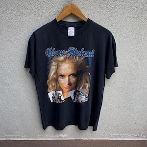 May include: Black t-shirt featuring a portrait of Gwen Stefani with her name in blue script above. The shirt includes two smaller images below the portrait. The shirt is hanging on a wooden hanger.