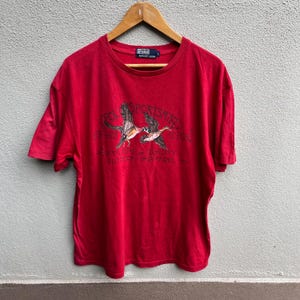 May include: Red short-sleeved t-shirt with a crew neck. The shirt features a graphic of two ducks in flight, with the words "POLO SPORTSMAN" and "SUPPLIERS OF QUALITY OUTDOOR APPAREL" printed on the front. The shirt is hanging on a wooden hanger.