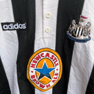 May include: Close-up of a black and white striped soccer jersey with the Adidas logo. The jersey features a Newcastle United crest and a "Newcastle Brown Ale" patch. The jersey has a white collar with buttons.