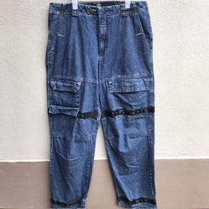 May include: Blue denim cargo trousers with multiple pockets and black accent straps. The trousers have a button and zip closure, with a relaxed fit. The black straps feature white text and cross symbols. The trousers are hanging on a white hanger.
