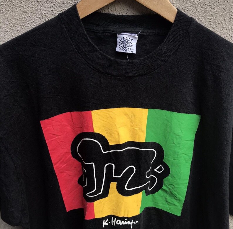 Puede incluir: Camiseta negra con un dise&ntilde;o gr&aacute;fico con bloques rojos, amarillos y verdes, y un contorno blanco de una figura que gatea. La camiseta tiene la firma "K. Haring" en la parte inferior.