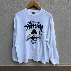 May include: White long-sleeve shirt featuring a black skull graphic and the word "Stussy". The design includes the text "Since 1980" and "USA". The shirt is displayed on a wooden hanger.