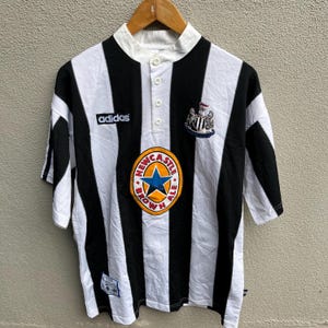 May include: A black and white striped soccer jersey with the Adidas logo on the chest. The jersey features a Newcastle Brown Ale logo and a team crest. The shirt has a button-up collar and short sleeves.