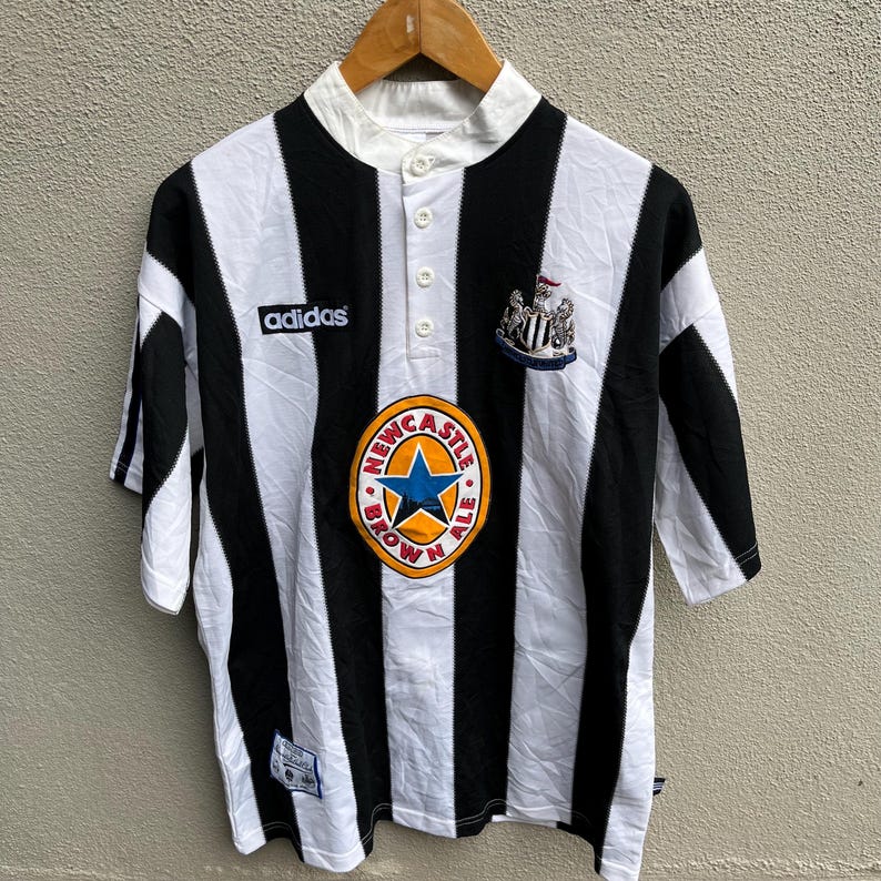 May include: A black and white striped soccer jersey with the Adidas logo on the chest. The jersey features a Newcastle Brown Ale logo and a team crest. The shirt has a button-up collar and short sleeves.
