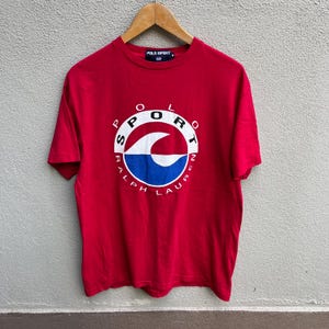 May include: A red Polo Sport t-shirt with a white and blue graphic design. The graphic features the word "SPORT" in a circular arrangement, with "RALPH LAUREN" around the bottom. The shirt is hanging on a wooden hanger.
