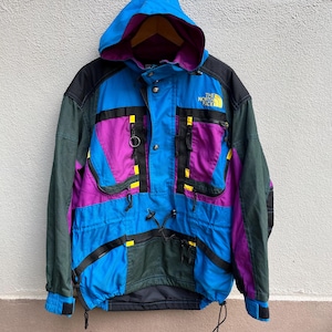 May include: A vintage color-blocked jacket with a hood. The jacket features a vibrant mix of blue, purple, black, and green panels, accented with yellow details. The North Face logo is visible on the chest. It has multiple pockets and drawstrings.