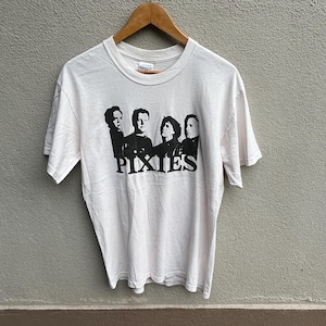 Puede incluir: Camiseta blanca con un estampado en blanco y negro de la banda Pixies. El gráfico muestra las caras de los miembros de la banda y el nombre de la banda en negrita, con una fuente sans-serif. La camiseta está colgada en una percha de madera.
