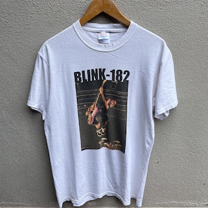 May include: White t-shirt featuring the band Blink-182. The graphic shows a person playing a pink guitar. The band's name is printed in black block letters above the image. The shirt has short sleeves and a crew neck.