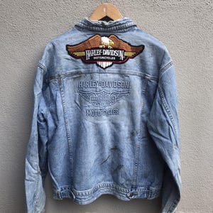 May include: Light blue denim jacket with Harley-Davidson Motorcycles logos embroidered on the back. The jacket features a large eagle emblem and the brand name in a classic font. The jacket is worn and has some stains.