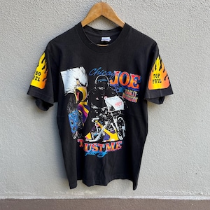 Puede incluir: Camiseta negra con gráfico de Harley-Davidson con un águila y un corredor de motos. La camiseta tiene el texto "Chicago Joe" y "Trust Me" con gráficos de llamas en las mangas. La camiseta está colgada en una percha de madera.
