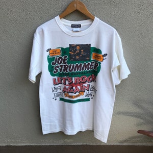 Może przedstawiać: Biała koszulka z graficznym wzorem dla Joe Strummer & The Mescaleros. Projekt zawiera tekst "Let's Rock Again" w kolorze czerwonym i zielonym, z dodatkowym tekstem i obrazami. Koszulka wisi na drewnianym wieszaku.
