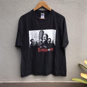 May include: Black t-shirt with a black and white graphic of The Simpsons family, and the word "Simpsons" in red. The t-shirt is displayed on a wooden hanger.