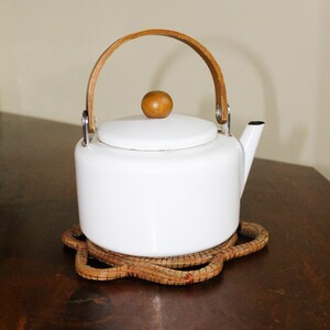 Könnte beinhalten: Weiße Email-Teekanne mit einem Holzgriff und -knopf. Die Teekanne steht auf einem geflochtenen braunen Untersetzer.