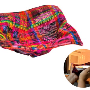 Peut inclure: Un bol en tissu tissé coloré, parfait pour servir des en-cas ou ranger de petits objets. Le bol est fait d'un tissu rouge vif, rose, jaune et bleu avec un motif à carreaux. Le bol est présenté en lévitation, avec une main tenant un bol similaire dans un four à micro-ondes en arrière-plan.