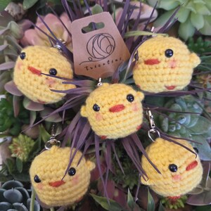 Peut inclure: Cinq porte-clés en forme de poussin jaune crocheté, avec des yeux noirs et des becs rouges. Chaque poussin a de petites ailes et un fermoir argenté. Une étiquette marron avec un logo et le texte "tricotini" est visible en arrière-plan.