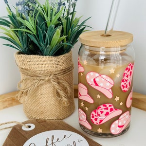 Puede incluir: Un vaso de vidrio con tapa de bamb&uacute; y pajita. El vaso tiene un fondo marr&oacute;n con un patr&oacute;n de sombrero de vaquero rosa y blanco. El texto "Coffee is always a good idea" est&aacute; en un letrero de madera en primer plano.