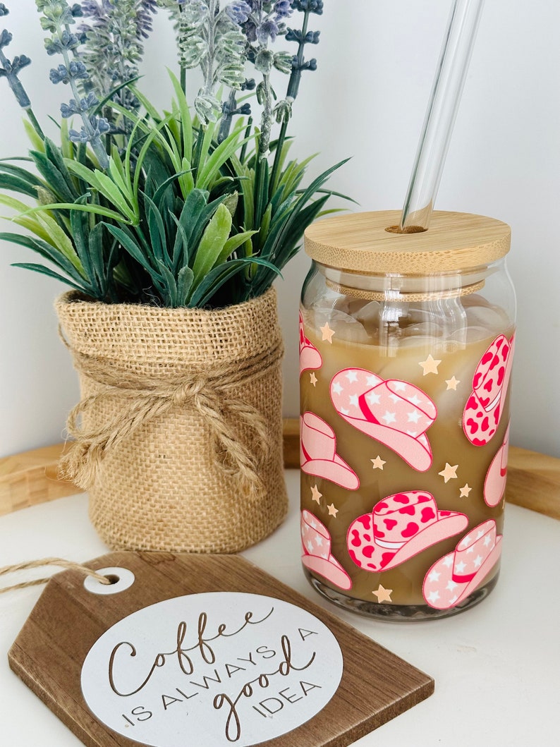 Puede incluir: Un vaso de vidrio con tapa de bamb&uacute; y pajita. El vaso tiene un fondo marr&oacute;n con un patr&oacute;n de sombrero de vaquero rosa y blanco. El texto "Coffee is always a good idea" est&aacute; en un letrero de madera en primer plano.