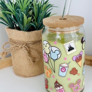 May include: A green glass jar with a bamboo lid and a glass straw. The jar is decorated with a cartoon design of Mickey Mouse, balloons, ice cream, and other Disney-themed imagery.
