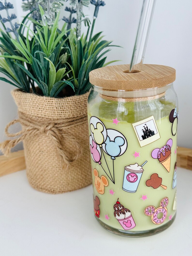 May include: A green glass jar with a bamboo lid and a glass straw. The jar is decorated with a cartoon design of Mickey Mouse, balloons, ice cream, and other Disney-themed imagery.