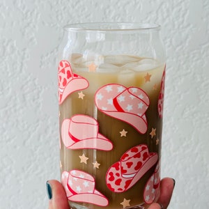 Puede incluir: Un vaso de vidrio transparente con una bebida marr&oacute;n y hielo. El vaso est&aacute; decorado con un patr&oacute;n de sombreros de vaquero rosas y rojos con estrellas blancas.