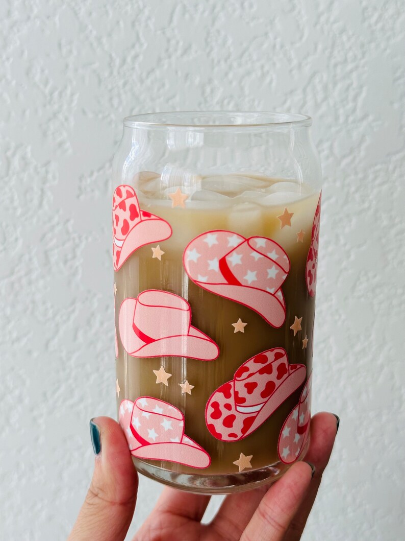 Puede incluir: Un vaso de vidrio transparente con una bebida marr&oacute;n y hielo. El vaso est&aacute; decorado con un patr&oacute;n de sombreros de vaquero rosas y rojos con estrellas blancas.