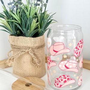 Puede incluir: Un vaso de vidrio transparente con un patr&oacute;n de sombrero de vaquero rosa y blanco. El vaso tiene una pajita y una tapa de bamb&uacute;.