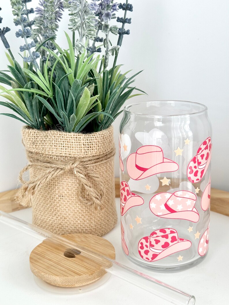 Puede incluir: Un vaso de vidrio transparente con un patr&oacute;n de sombrero de vaquero rosa y blanco. El vaso tiene una pajita y una tapa de bamb&uacute;.