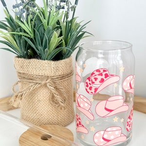 Puede incluir: Un vaso de vidrio transparente con un dise&ntilde;o de sombrero de vaquero rosa y blanco. El vaso tiene una pajita y un posavasos de madera.