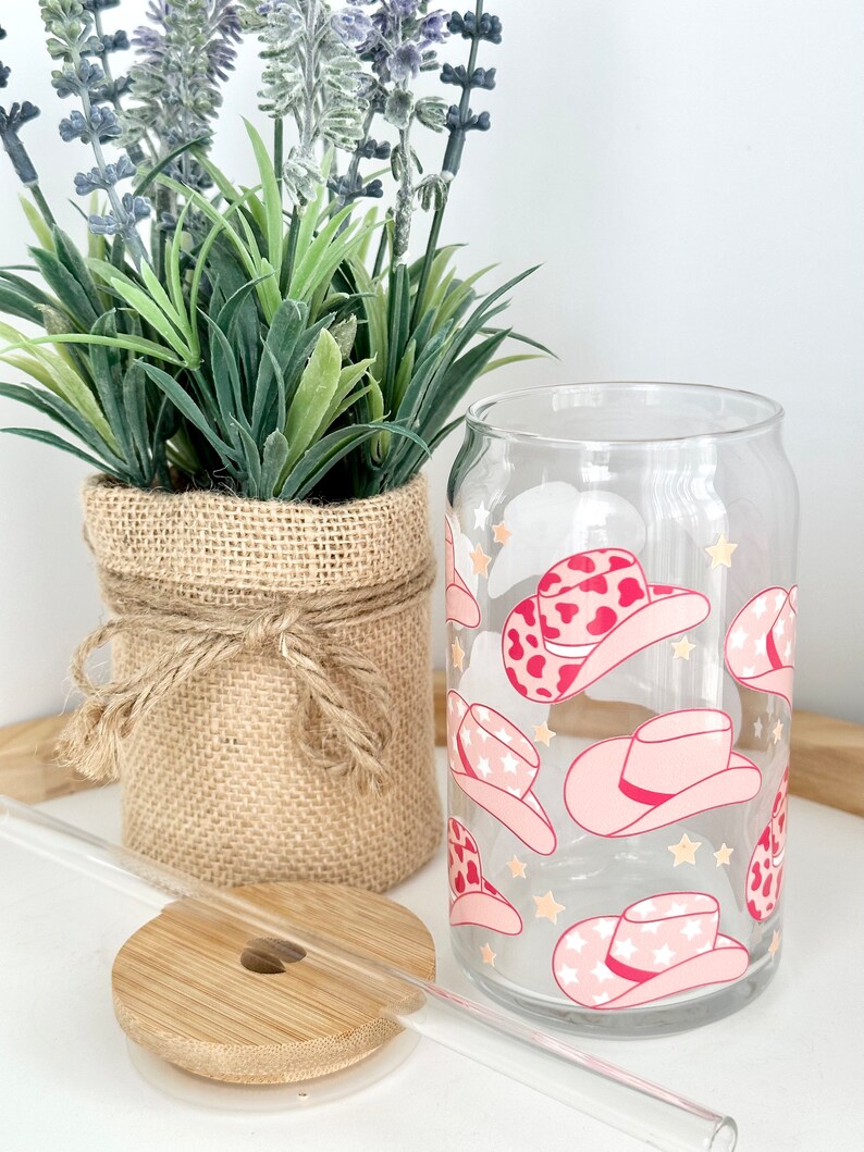 Puede incluir: Un vaso de vidrio transparente con un dise&ntilde;o de sombrero de vaquero rosa y blanco. El vaso tiene una pajita y un posavasos de madera.