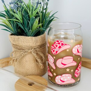 Puede incluir: Un vaso de vidrio transparente con un patr&oacute;n de sombrero de vaquero marr&oacute;n y rosa. El vaso est&aacute; lleno de caf&eacute; helado y tiene una pajita al lado. El vaso est&aacute; sobre una posavasos de madera.