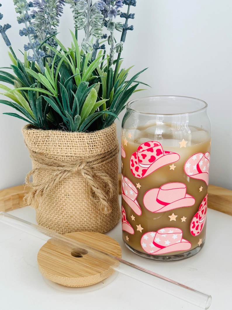 Puede incluir: Un vaso de vidrio transparente con un patr&oacute;n de sombrero de vaquero marr&oacute;n y rosa. El vaso est&aacute; lleno de caf&eacute; helado y tiene una pajita al lado. El vaso est&aacute; sobre una posavasos de madera.