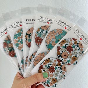 May include: Set of six floral car coasters with a white background and a variety of colors, including red, orange, yellow, green, and blue. The coasters are packaged in clear plastic sleeves with the text "Car Coasters Handmade with love" printed on them.