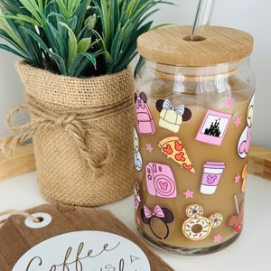 May include: A glass jar with a bamboo lid and a straw. The jar is decorated with pink and white stickers featuring Disney characters and icons. The jar is filled with a brown liquid and ice. The text "Coffee is always a good idea" is on a wooden sign in the foreground.