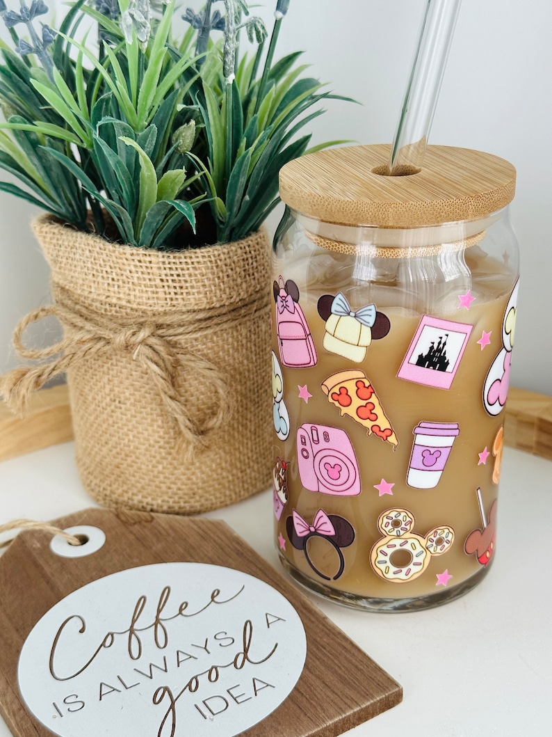 May include: A glass jar with a bamboo lid and a straw. The jar is decorated with pink and white stickers featuring Disney characters and icons. The jar is filled with a brown liquid and ice. The text "Coffee is always a good idea" is on a wooden sign in the foreground.