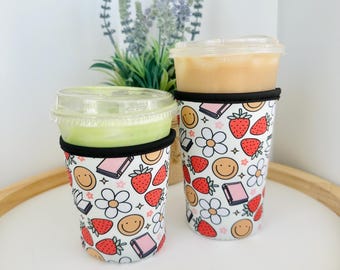 May include: Two reusable beverage sleeves with a white background and a colorful pattern of strawberries, flowers, books, and smiley faces. The sleeves are made of neoprene and are designed to fit most standard-sized cups.