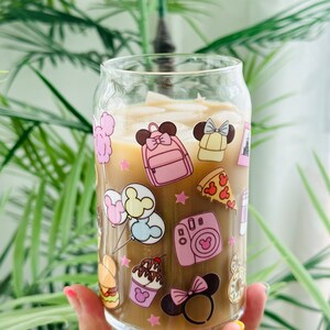 May include: A clear glass tumbler with a brown beverage inside. The tumbler has a colorful design featuring cartoon images of Mickey Mouse, Minnie Mouse, and other Disney characters. The design includes images of food, balloons, and a camera.