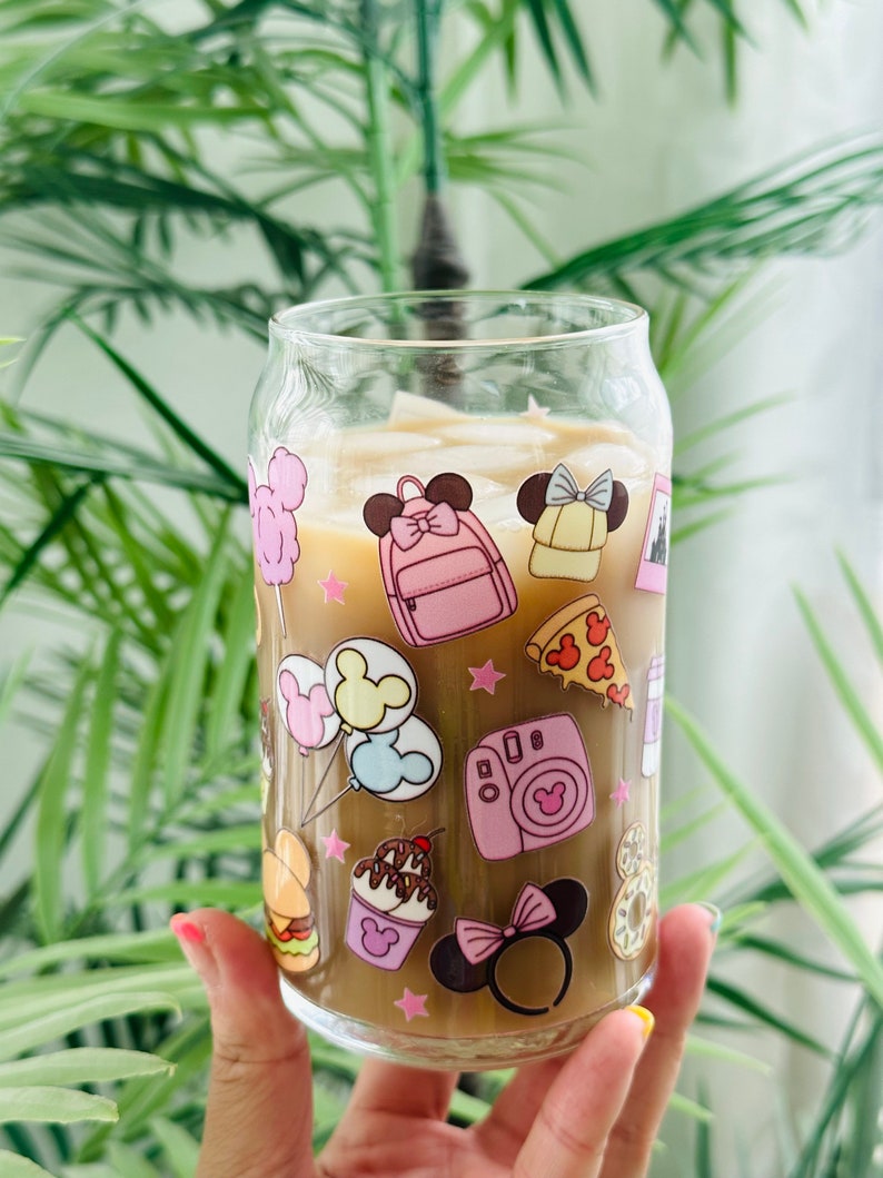 May include: A clear glass tumbler with a brown beverage inside. The tumbler has a colorful design featuring cartoon images of Mickey Mouse, Minnie Mouse, and other Disney characters. The design includes images of food, balloons, and a camera.
