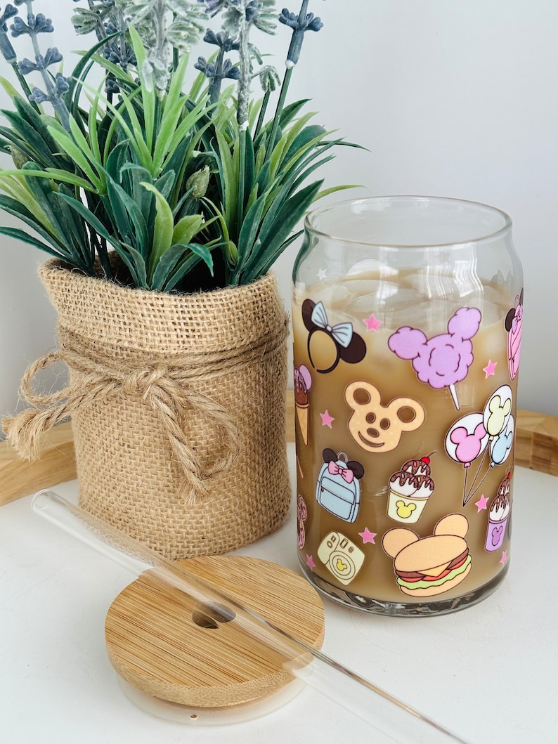 May include: A clear glass can with a straw and a bamboo lid. The can has a cartoon design featuring a mouse character, ice cream, and other Disney-themed imagery.