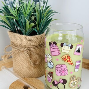 May include: A clear glass tumbler with a green background and pink and white cartoon images of Mickey Mouse and Minnie Mouse. The tumbler has a straw and a wooden lid.