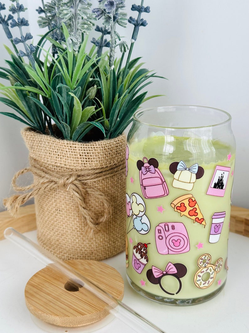 May include: A clear glass tumbler with a green background and pink and white cartoon images of Mickey Mouse and Minnie Mouse. The tumbler has a straw and a wooden lid.