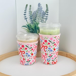 May include: Two reusable coffee cup sleeves with floral and bow patterns. The sleeves are white with red bows, pink and red flowers, and blue flowers. The sleeves are around clear plastic cups with drinks inside.