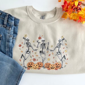 May include: A beige crewneck sweatshirt featuring a graphic print of three dancing skeletons surrounded by orange pumpkins, yellow sunflowers, and scattered autumn leaves in shades of red, orange, and yellow, paired with blue denim jeans.