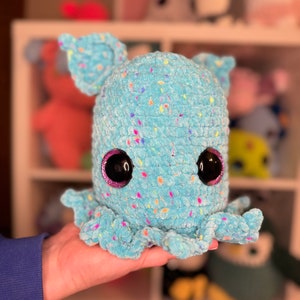 Puede incluir: Un pulpo de crochet azul con motas de colores y grandes ojos negros. El pulpo está siendo sostenido en la mano de una persona.